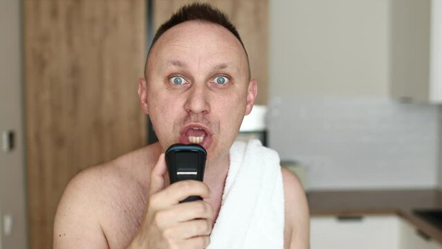 young unshaven man with towel on shoulders shaving with modern electric razor trimer while looking at camera, indoors. Daily routine male self-care. man shaves with an electric razor in the kitchen.