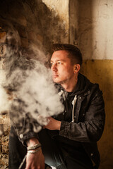 Portrait of a brutal smoking macho man in a black denim jacket with a stylish hairstyle, piercing in the eyebrow and lip and an earring in the ear. A rock musician or punk in an abandoned building