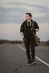 Portrait of a brutal macho man in a black denim jacket with a stylish hairstyle, piercing in the eyebrow and lip and an earring in his ear, walking along the road with a bag on his shoulder