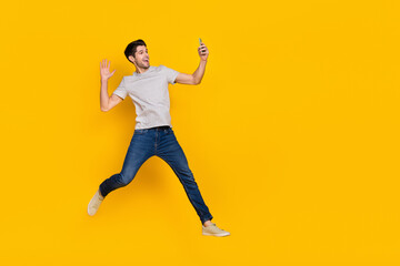 Fototapeta na wymiar Full size photo of funny brunet millennial guy jump do selfie wear t-shirt jeans shoes isolated on yellow background