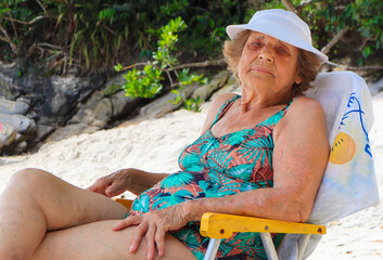 Vovó idosa, sentada na cadeira de praia, usando chapéu branco e maio colorido, tomando Sol e relaxando na praia - obrazy, fototapety, plakaty