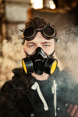 A brutal guy with an eyebrow piercing in cyberpunk or steampunk goggles on his forehead and a respirator on his face amid smoke in an overloaded building. Apocalypse male portrait