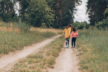 Fototapeta premium Little girl friends walk holding hands along a rural path near the forest, summer vacation