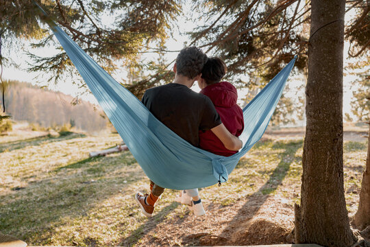 Rear of woman and man sitting in hammock and hugging while having rest in forest. Relaxation.