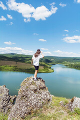 Naklejka na ściany i meble Young beautiful traveler woman sitting on a rock above lake .Traveling in the summer nature
