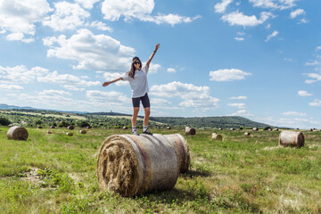 Naklejka na ściany i meble Happy traveler woman standing on haystacks on blue cloudy sky background .Travel in Bulgaria Concept 