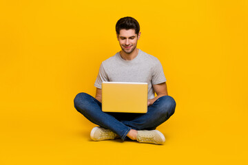 Full size photo of nice brunet young guy sit type laptop wear t-shirt jeans footwear isolated on yellow background