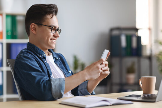Millennial Businessman Using Smartphone While Sitting At Desk In Office
