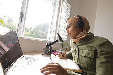 Smiling biracial young woman in hijab recording podcast through microphone at home studio