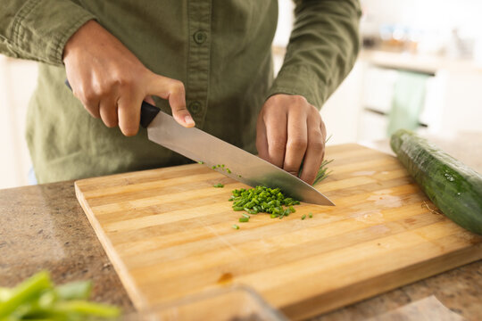 Midsection of biracial young woman cutting scallions on wooden board in kitchen at home