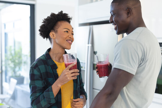 Happy african american mid adult couple holding smoothie in glasses looking at each other in kitchen