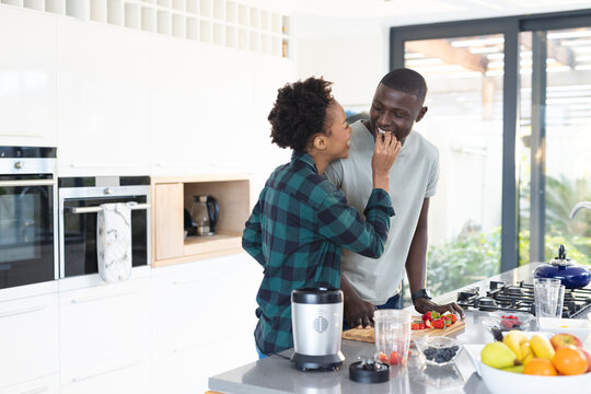 African american happy mid adult woman feeding berry to boyfriend while standing at kitchen island