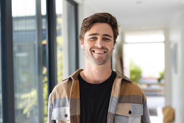 Portrait of smiling caucasian young man wearing jacket standing by window at home