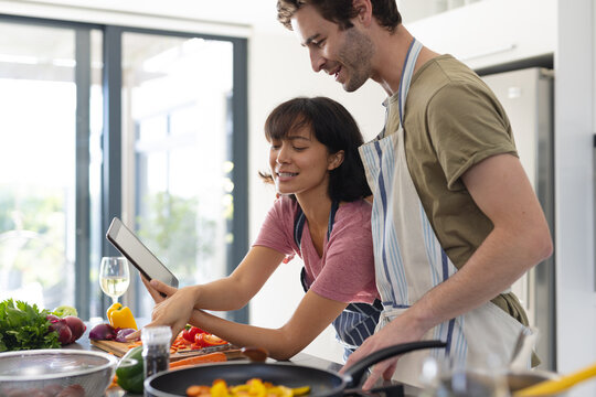 Multiracial young couple watching recipe over digital tablet while cooking food on kitchen island