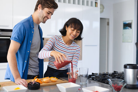 Smiling caucasian boyfriend looking at asian girlfriend pouring fruits smoothie in glass in kitchen