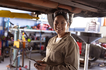 Portrait of smiling mid adult asian mechanic repairing car using digital tablet in workshop