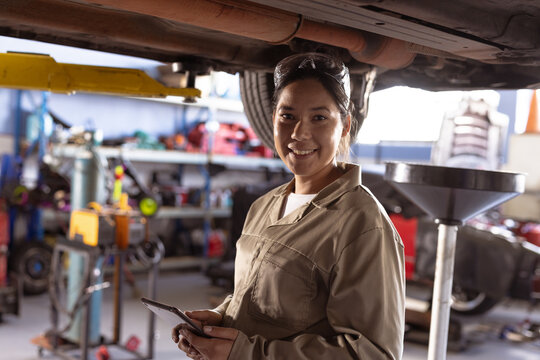 Portrait of smiling mid adult asian mechanic repairing car using digital tablet in workshop - Powered by Adobe