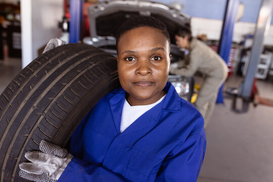 Portrait of confident female mid adult african american mechanic carrying car's tire in workshop