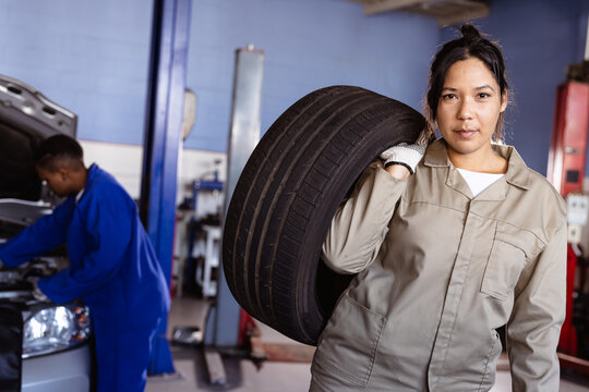 Portrait of confident mid adult asian female mechanic carrying car's tire in workshop, copy space