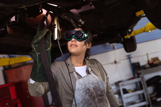 Mid adult asian female welder using work tools while wielding car under car lift in workshop