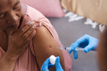 Cropped hands of african american doctor injecting medicine on senior woman's shoulder at home