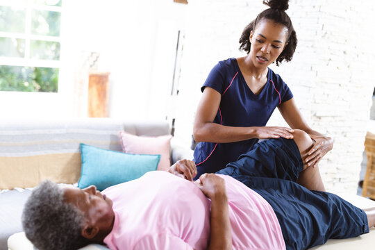 African american female physiotherapist massaging senior woman's leg lying on table at home