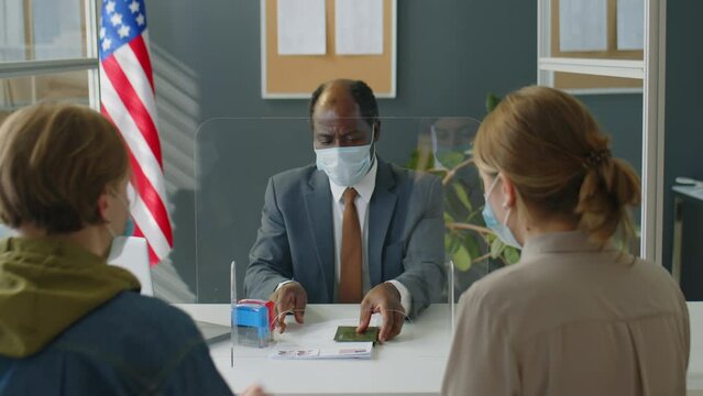 African American consular officer in protective face mask sitting behind glass screen and interviewing couple of visa applicants while working in US embassy during coronavirus outbreak