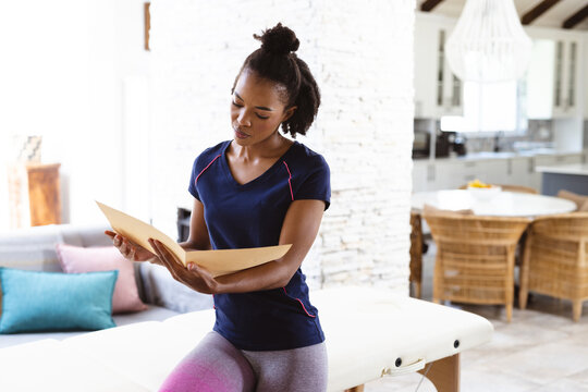 African american female physiotherapist reading medical report while sitting on table at home