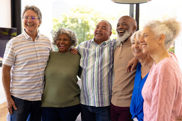 Multiracial senior friends with arms around laughing and looking away while standing in nursing home
