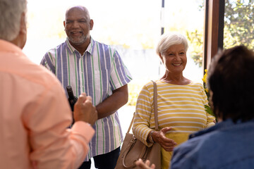 Biracial seniors welcoming cheerful friends standing at doorway in nursing home, copy space