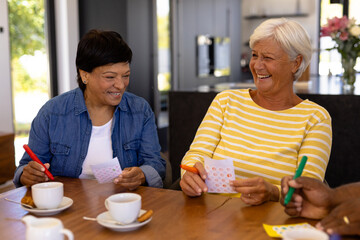 Cheerful biracial senior female friends with coffee and cookies on dining table playing bingo