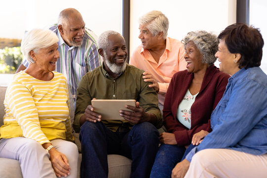 Multiracial Senior Man Holding Digital Tablet Looking At Laughing Friends While Sitting On Sofa