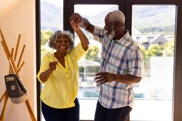 Cheerful african american senior man doing salsa dance with woman against window in nursing home