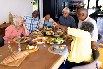 Fototapeta na wymiar Multiracial senior woman showing smart phone to friends while having lunch at dining table