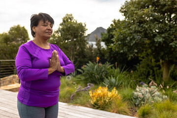 Biracial senior woman with eyes closed meditating while standing against plants in yard