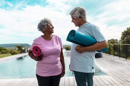 Happy Multiracial Senior Friends Holding Yoga Mats Talking While Standing At Poolside Against Sky