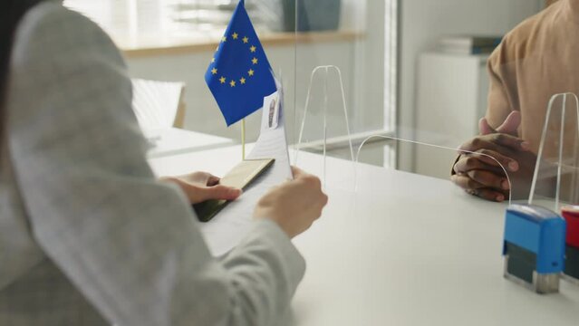 African American man in mask giving application form and passport to consular officer while sitting behind glass shield during schengen visa interview in EU embassy in times of pandemic