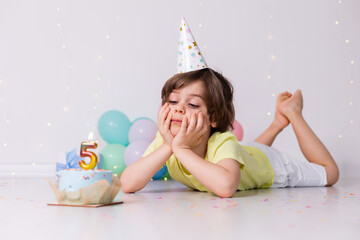 Fototapeta premium Cute little birthday boy in hat, cake with candle five, balloons, happy birthday