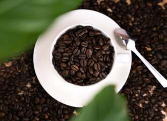 White cup with fresh fragrant coffee beans and green leaves in defocus. Top view. Selective focus.
