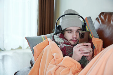 Heating season. Male teenager is freezing, covered with warm blanket, sitting on the couch at cold...