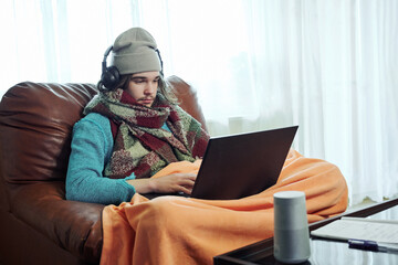 Cold season, frozen teenager in warm cap under blanket sitting near a heating radiator with laptop...