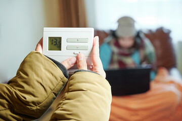 Indoor thermometer with low temperature of air in female hands. Frozen teenager in warm cap is...