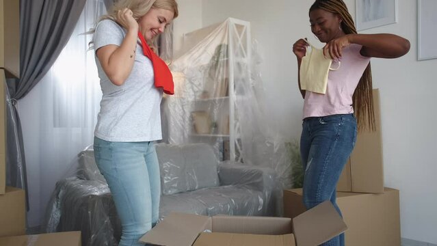 Happy relocation. New home. Real estate mortgage. Satisfied multiethnic female girls moving together unpacking boxes with clothes stuff in apartment.