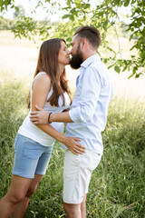 Fototapeta premium pretty young pregnant woman with white shirt stands with her boyfriend with beard and blue shirt in high flower meadow and cuddle