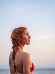Naklejka premium Portrait of a fair-haired girl in a bikini standing on the background of the sea on a summer day. Profile view