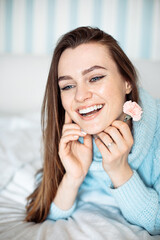 Romantic young woman with toothy smile in blue sweater lying on bed with flower in hand. Portrait...