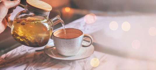 Banner of grey cup with teapot. The hand holding the teapot pours herbal tea into pot on saucer in soft focus on blurred background. Coffee, tea house, bokeh lights. The concept of cozy pastime - Powered by Adobe