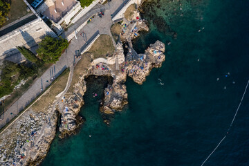 Fototapeta na wymiar Top aerial view of a rock beach with beautiful blue green water in Rovinj on the Mediterranean sea with road and buildings on the coast