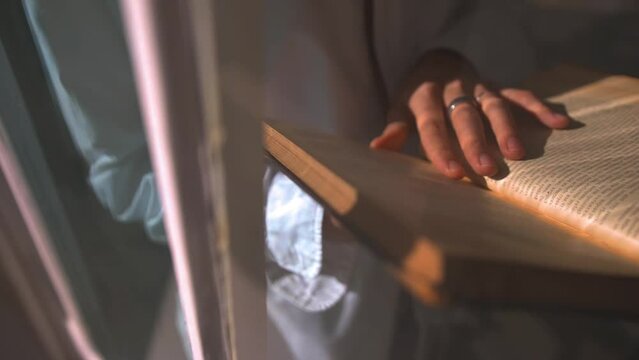 Young man reading a vintage book on a windowsill. Guy touches aged book pages to feel the texture of a paper. Dissertation writing on an ancient volume of a rare edition of a book, education process.
