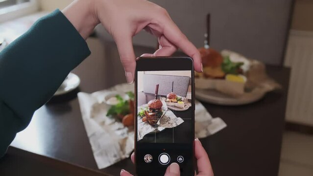A woman takes a picture on a smartphone of a delicious burger on a table in a cafe for social networks.